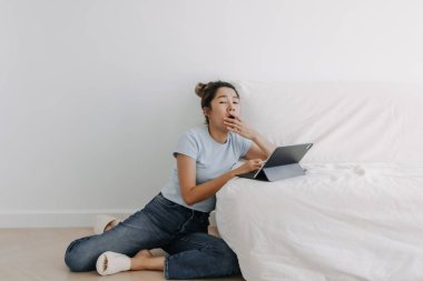 Asyalı genç Taylandlı kadın, beyaz kanepeye yaslanmış, apartman odasında tablet kullanıyor, uykulu ve bilgisayarla tek başına çalışırken esniyor..