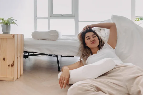 Asian Thai woman falling down from bed, sitting on floor and leaning the bed, lazy relaxing alone in home apartment.