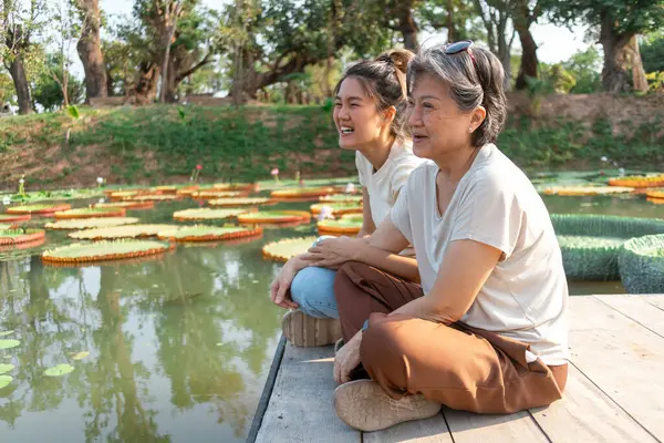 Side view of asian Thai Chinese elder mother and daughter sitting on the bank by lotus flower lake local lagoon, both woman happy smiling, travel on vacation under the sun.