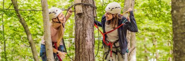 BANNER, BANNER, LONG FORMAT İki kadın kız ormanda, ağaçtaki karabina emniyet halatıyla, ağaç tepesindeki halat macera parkına doğru süzülerek ilerliyorlar. Aile hafta sonu çocukları.