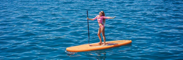 バンナー ロングフォーマット若い女性はモンテネグロの青い海でパドリングを楽しんでいます Sup 岩の近くのパドルボード上の女の子のトレーニング — ストック写真