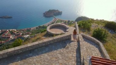 Genç kadın, Budva kenti yakınlarındaki Sveti Stefan adasının üzerindeki Saint Sava kilisesini ziyaret ediyor. O, bakış açısına doğru koşuyor. Karadağ 'a seyahat. Yavaş çekim hava görüntüsü.