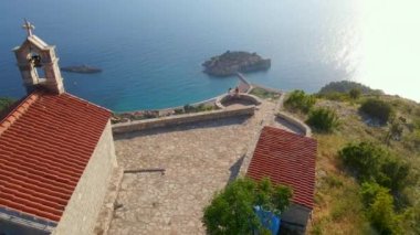 Havadan yavaş çekim videosu. Budva kenti yakınlarındaki Sveti Stefan adasının üzerindeki Saint Sava kilisesini iki kadın ziyaret ediyor. Karadağ 'a Seyahat.