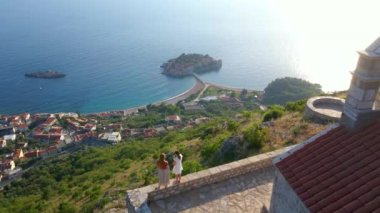 Budva kenti yakınlarındaki Sveti Stefan adasının üzerindeki Saint Sava kilisesini iki kadın ziyaret ediyor. Karadağ 'a seyahat. Yavaş çekim videosu.