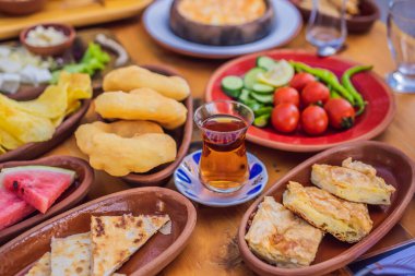 Türk kahvaltı masası. Hamur işleri. Sebzeler. Zeytin. Peynir, yağda yumurta. Reçel, bakır demlikte çay ve lale bardakları. Geniş bileşim.