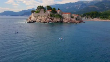 Yavaş çekim hava görüntüsü. Budva yakınlarındaki Sveti Stefan adasının etrafında SUP kürek çeken genç bir kadın. Karadağ 'da yaz.