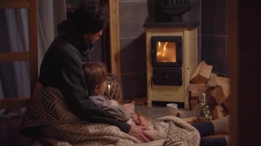 Genç bir adam ve küçük oğlu battaniyeye sarılı bir sobanın başında oturup ateşe bakıyorlar. Küçük sıcacık ahşap ev. Kış tatili konsepti.