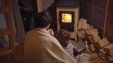 Genç bir adam ve küçük oğlu battaniyeye sarılı bir sobanın başında oturup ateşe bakıyorlar. Küçük sıcacık ahşap ev. Kış tatili konsepti.