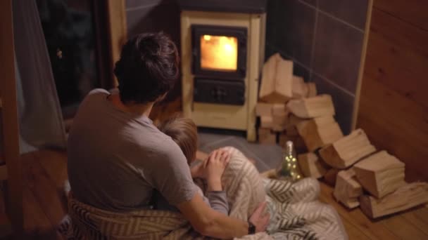 若い男と彼の小さな息子が毛布の中に火を見て 家庭用ストーブで座って包まれた 小さな居心地の良い木造住宅 冬休みのコンセプト — ストック動画