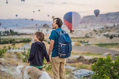 Kapadokya, Türkiye 'de sıcak hava balonlarına bakan baba ve oğul turistler. İyi yolculuklar hindi konsepti. Baba ve oğul dağın tepesinde harika manzaranın tadını çıkarıyorlar..