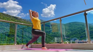 Genç bir kadının yavaş çekim videosu, arka planda dağlar ve deniz olan modern apartman terasında yoga yapıyor..