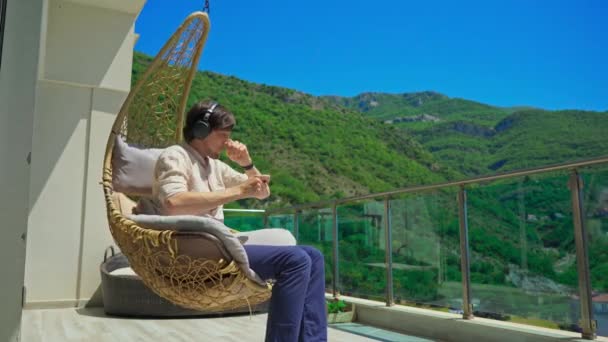 Young Man Balcony Enjoying Music Swinging Chair Mountain Scenery Background — Stockvideo