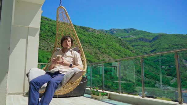 年轻人在阳台上休息 喝咖啡 四周环绕着山水 在一个舒适的摇椅里 在宁静的环境中 理想的休闲 旅行和休闲环境 — 图库视频影像