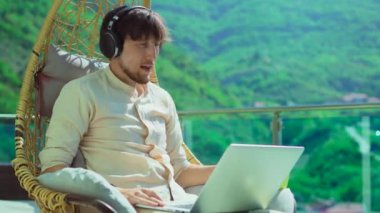 Young man on balcony, surrounded by stunning mountain scenery. In a comfortable swinging chair, he wears headphones and talks on a laptop while drinking coffee or tea. He disagrees with something