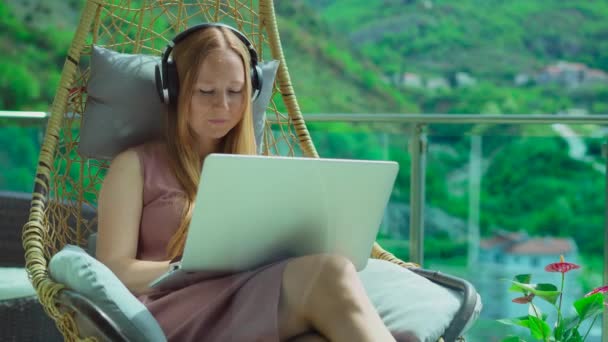 阳台上的年轻女子 四周环绕着令人惊叹的山景 舒适地坐在摇椅上 她戴着耳机 键盘上打字 完美的展示技术 通信或 — 图库视频影像