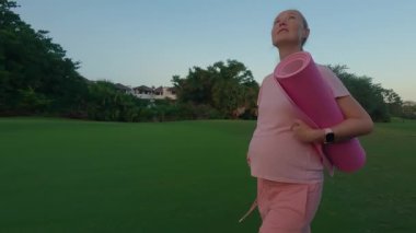 A stunning slow-motion stock video captures a beautiful moment of motherhood. A pregnant woman, dressed in a pink t-shirt and shorts, walks gracefully through a lush green field with a yoga mat in