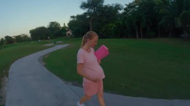 A stunning slow-motion stock video captures a beautiful moment of motherhood. A pregnant woman, dressed in a pink t-shirt and shorts, walks gracefully through a lush green field with a yoga mat in