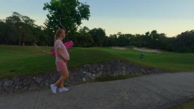 A stunning slow-motion stock video captures a beautiful moment of motherhood. A pregnant woman, dressed in a pink t-shirt and shorts, walks gracefully through a lush green field with a yoga mat in