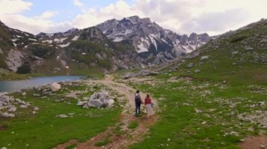 Aerial video. A young couple, dressed in casual clothes, takes a leisurely stroll through a lush valley with glacier lake and snow-capped mountains in the background. The peaceful surroundings set the