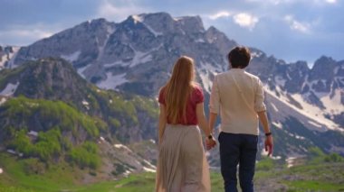 A young couple stands in a beautiful valley surrounded by snow-capped mountains, holding hands and gazing out at the stunning landscape around them. Dressed casually, as if on a carefree road trip