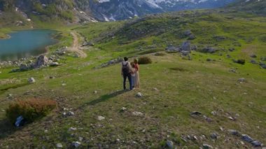 A young family captures their outdoor adventure using a drone camera. The father, mother, and their son stand in a breathtaking mountain valley with snow-capped peaks in the background, dressed