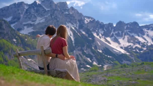 一对身穿休闲装的年轻夫妇坐在一张木制长椅上 凝视着外面一个风景如画的山谷 后面是白雪覆盖的高山 柔和的微风和温暖的阳光带来了 — 图库视频影像