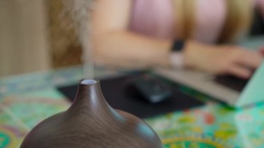 The video features a young woman sitting at a desk in her apartment, working on her laptop. A working electric aromatherapy diffuser is seen to the side of her, emitting a soothing fragrance. The