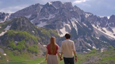 A young couple, dressed in casual clothes, takes a leisurely stroll through a lush valley with snow-capped mountains in the background. The peaceful surroundings set the tone for the couples relaxed