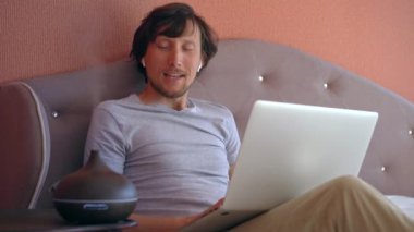 A young woman is seen sitting comfortably on a bed, working on his laptop while a working electric aroma diffuser is placed in front of him. The diffuser releases a soothing fragrance into the air