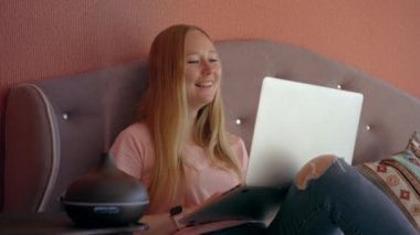 A young woman is seen sitting comfortably on a bed, working on her laptop while a working electric aroma diffuser is placed in front of her. The diffuser releases a soothing fragrance into the air