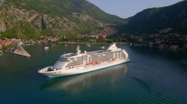 Yavaş çekim videosu. Güneş Boka-Kotor Körfezi 'nde batarken, limandan ayrılan beyaz bir yolcu gemisi arkasında hafif dalgalar bırakır. Dağların göz kamaştırıcı manzarası ve eski kasaba
