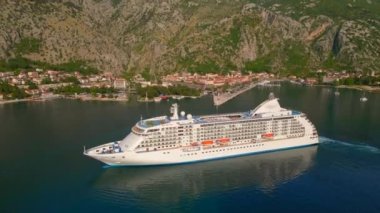 Yavaş çekim videosu. Güneş Boka-Kotor Körfezi 'nde batarken, limandan ayrılan beyaz bir yolcu gemisi arkasında hafif dalgalar bırakır. Dağların göz kamaştırıcı manzarası ve eski kasaba