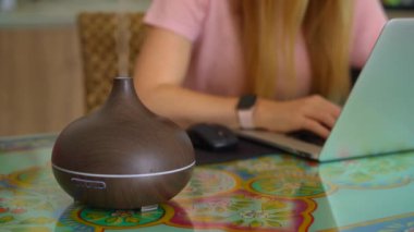 The video features a young woman sitting at a desk in her apartment, working on her laptop. A working electric aromatherapy diffuser is seen to the side of her, emitting a soothing fragrance. The
