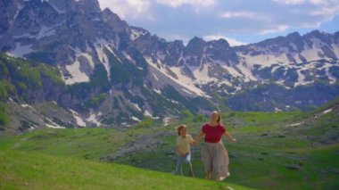 Yavaş çekim videosu. Gündelik giysiler içinde genç bir kadın ve oğlu arka planda karla kaplı dağlarla dolu yemyeşil bir vadide koşuyorlar. Bu görüntü projeler için mükemmel.