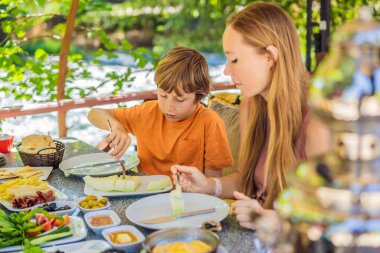 Anne ve oğlu Türk kahvaltısı yapıyor. Türk kahvaltı masası. Hamur işleri. Sebzeler. Zeytin. Peynir, yağda yumurta. Reçel, bakır demlikte çay ve lale bardakları. Geniş bileşim.