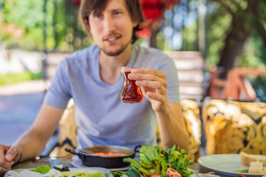 Erkek turist Türk kahvaltısı yapar. Türk kahvaltı masası. Hamur işleri. Sebzeler. Zeytin. Peynir, yağda yumurta. Reçel, bakır demlikte çay ve lale bardakları. Geniş bileşim.