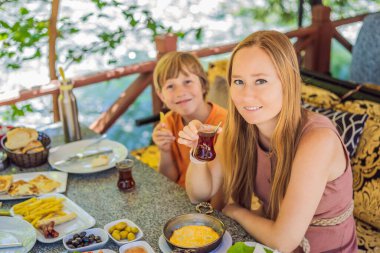 Anne ve oğlu Türk kahvaltısı yapıyor. Türk kahvaltı masası. Hamur işleri. Sebzeler. Zeytin. Peynir, yağda yumurta. Reçel, bakır demlikte çay ve lale bardakları. Geniş bileşim.