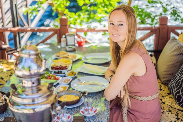 woman tourist eat Turkish breakfast. Turkish breakfast table. Pastries. Vegetables. Olives. Cheeses, fried eggs. Jams, tea in copper pot and tulip glasses. Wide composition.