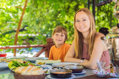 Anne ve oğlu Türk kahvaltısı yapıyor. Türk kahvaltı masası. Hamur işleri. Sebzeler. Zeytin. Peynir, yağda yumurta. Reçel, bakır demlikte çay ve lale bardakları. Geniş bileşim.