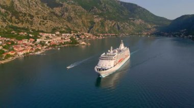 Yavaş çekim videosu. Güneş Boka-Kotor Körfezi 'nde batarken, limandan ayrılan beyaz bir yolcu gemisi arkasında hafif dalgalar bırakır. Dağların göz kamaştırıcı manzarası ve eski kasaba