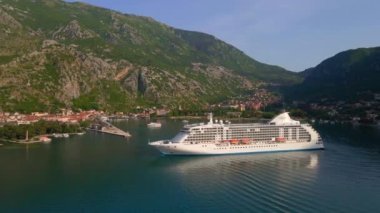 Yavaş çekim videosu. Güneş Boka-Kotor Körfezi 'nde batarken, limandan ayrılan beyaz bir yolcu gemisi arkasında hafif dalgalar bırakır. Dağların göz kamaştırıcı manzarası ve eski kasaba
