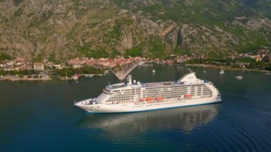 Yavaş çekim videosu. Güneş Boka-Kotor Körfezi 'nde batarken, limandan ayrılan beyaz bir yolcu gemisi arkasında hafif dalgalar bırakır. Dağların göz kamaştırıcı manzarası ve eski kasaba