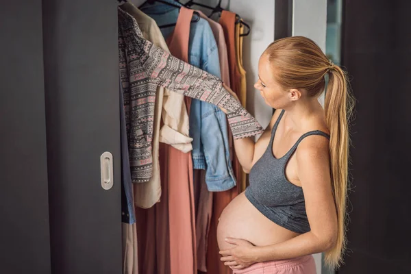 A pregnant woman has nothing to wear. A pregnant woman stands in front of a closet with clothes and does not know what to wear because the clothes do not fit on her.