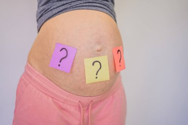 Bebek ismi seçmek. Karındaki kağıt çıkartmalarında soru işaretleri olan kafası karışmış hamile bir kadın..