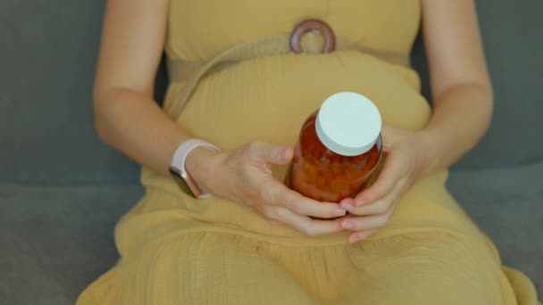 このスローモーションビデオでは 妊娠中の女性がソファに座っている間にビタミンや食品サプリメントの瓶を持っているのが見られます 彼女は瓶を開けて手の手のひらにいくつかの薬を置く — ストック動画