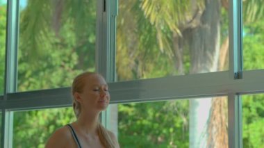 Bu yavaş çekim videosunda, genç hamile bir kadın dairesinde panoramik bir pencere ve arka planda yemyeşil bir tropikal zemin ile yoga egzersizleri yaparken görülebilir. Yavaş ve...