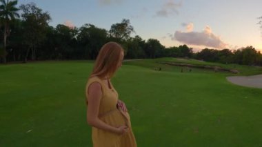 Bu büyüleyici yavaş çekim videosunda, hamile bir kadın canlı yeşil bir alanda zarif bir şekilde yürüyor, güzel bir günbatımının büyüleyici renkleriyle aydınlatılıyor. Sakin atmosfer ve kadınlar.