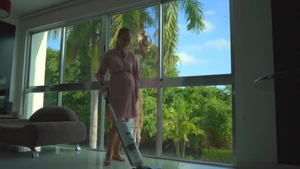 ピンクのドレスを着た妊婦が コードレスのない電気モップの掃除機で床をきれいに掃除する様子がストック動画で紹介されています カメラは彼女の立ち位置をパノラマの窓で捉える — ストック動画