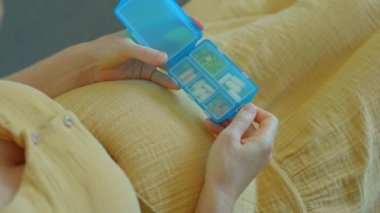 Bu yavaş çekim videosunda, hamile bir kadın kanepede otururken görülüyor. Parçalı plastik bir kutudan vitamin ya da gıda takviyesi alıyor. Kamera onun ellerine odaklanmış durumda.