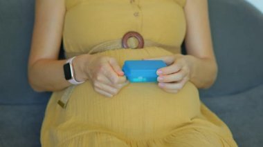 Bu yavaş çekim videosunda, hamile bir kadın kanepede otururken görülüyor. Parçalı plastik bir kutudan vitamin ya da gıda takviyesi alıyor. Kamera onun ellerine odaklanmış durumda.